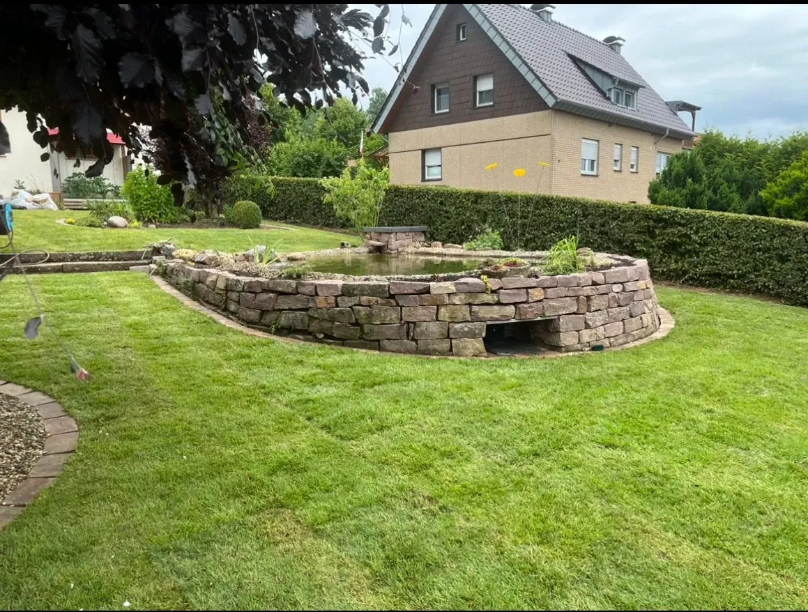 Ein üppiger grüner Rasen mit einer Steinmauer und einem Teich vor einem Haus.