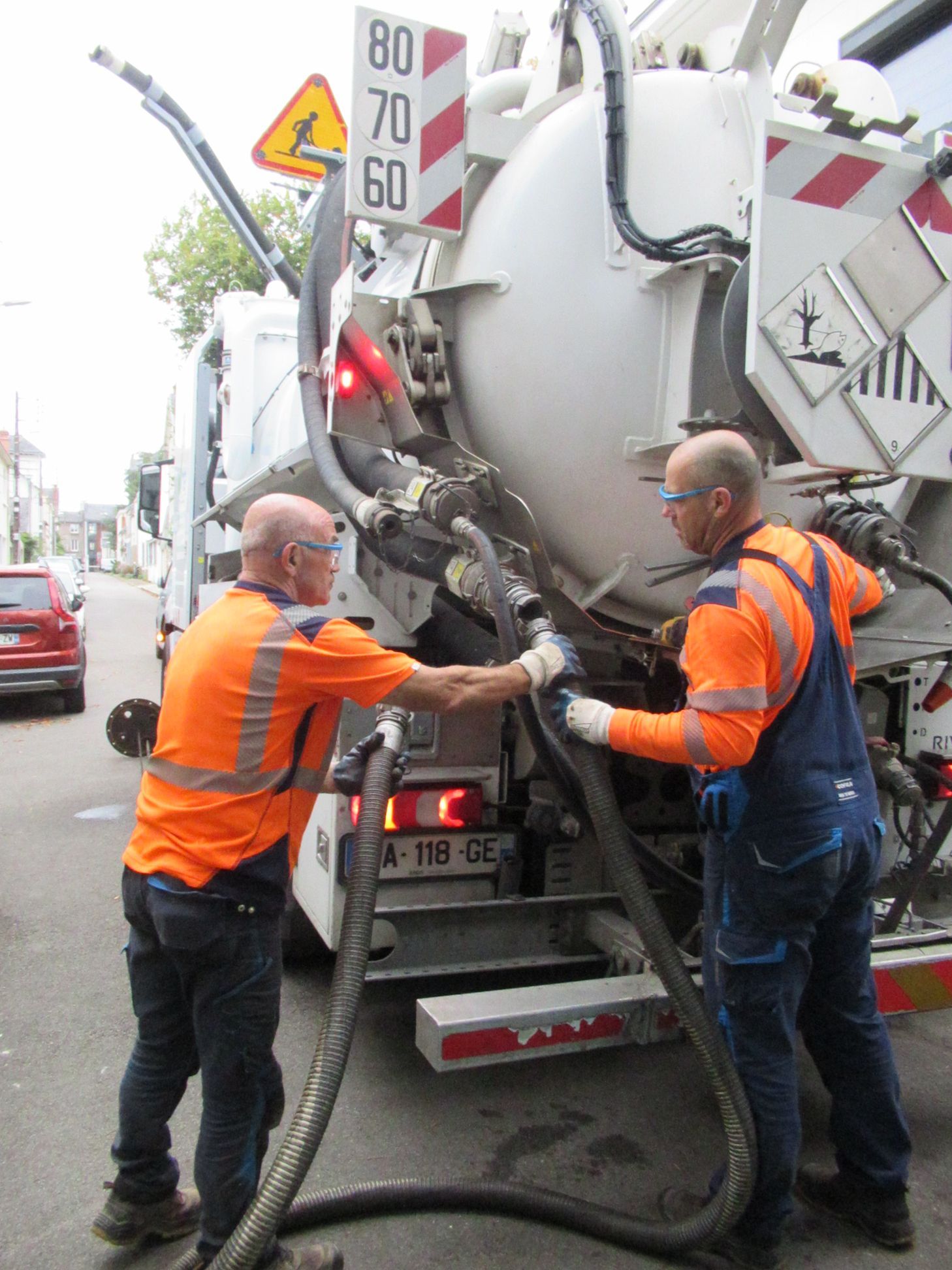 Deux ouvriers en gilet orange manipulent des tuyaux près d’un camion malaxeur à béton dans une rue.