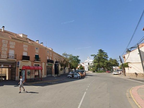 Una calle de un pueblo con edificios, coches aparcados y una persona caminando. El cielo es azul.
