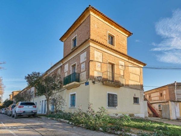 Edificio de ladrillo de varias plantas con torre, ubicado en una calle con coches aparcados bajo un cielo azul. Se ven algunas ventanas y balcones.