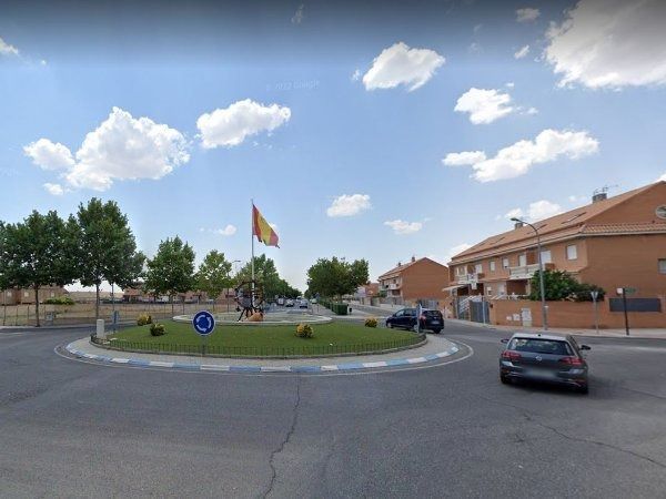 Una rotonda en una plaza de la ciudad con una bandera española, coches y edificios residenciales bajo un cielo nublado.