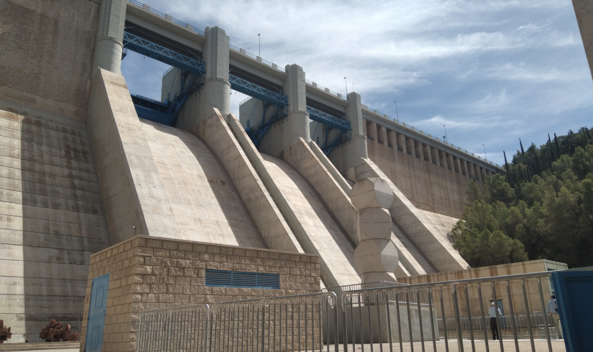 Una presa de hormigón con compuertas de liberación de agua, situada frente a una ladera boscosa bajo un cielo parcialmente nublado.