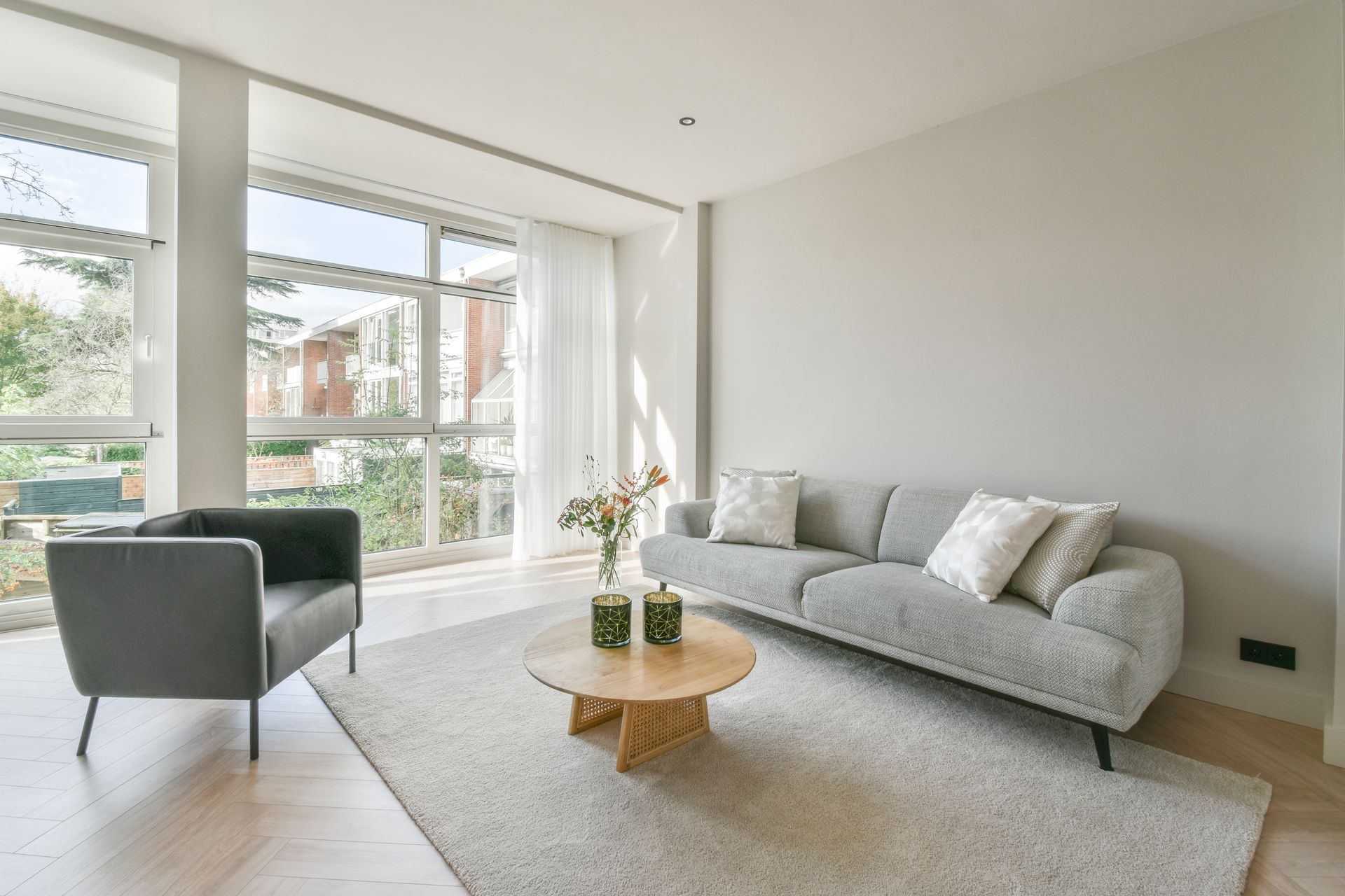 Salon avec un canapé gris, un fauteuil devant de grandes fenêtres.