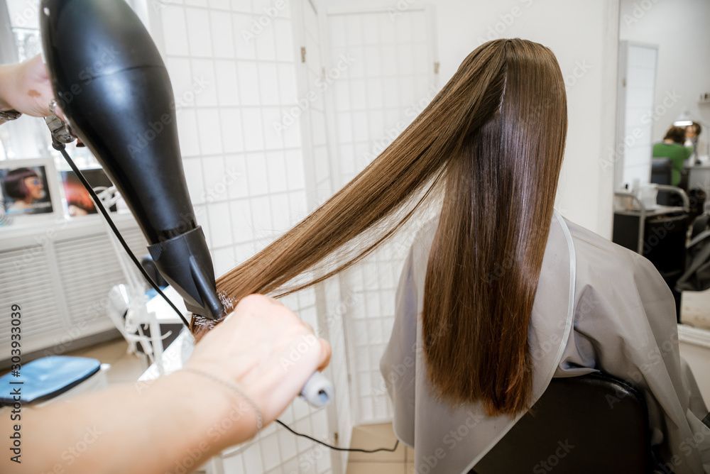 Haare werden im Friseurstuhl mit einem schwarzen Haartrockner geföhnt, langes braunes Haar ist ausgebreitet.