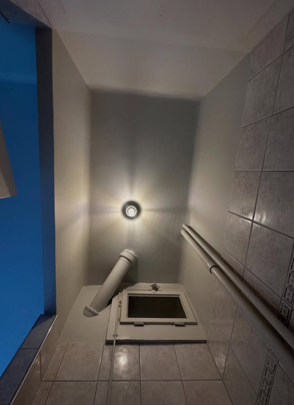 Pièce étroite carrelée avec une porte à trappe, des tuyaux et un plafonnier lumineux.