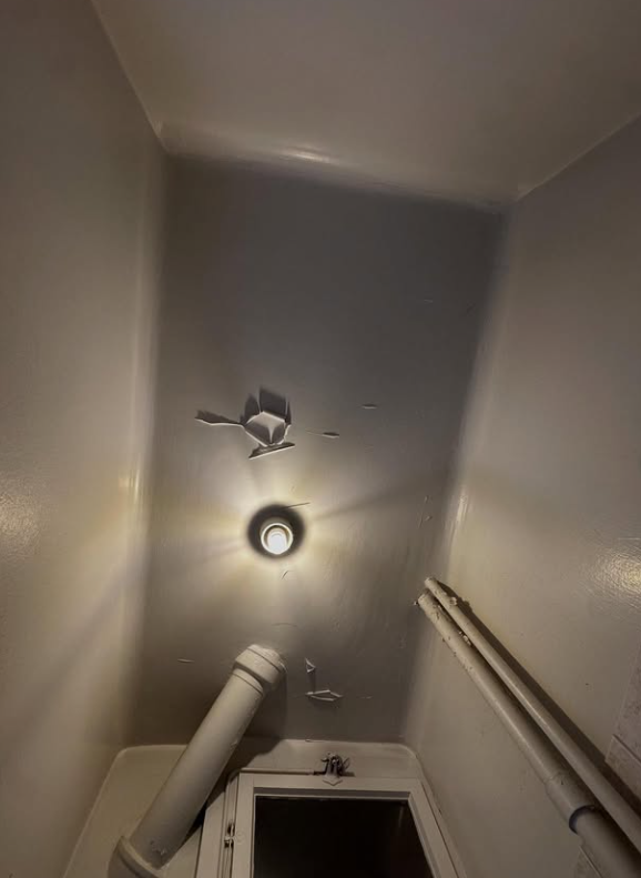 Vue en contre-plongée d'une pièce grise au plafond endommagé. Un luminaire est allumé et un tuyau est visible.