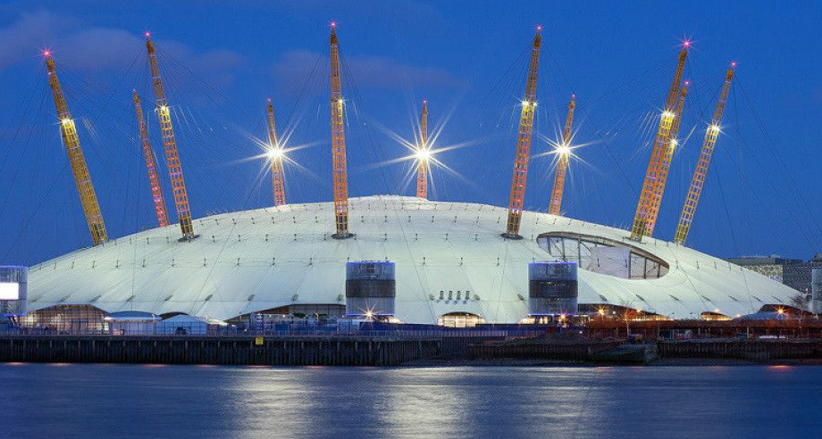 The O2 arena in London illuminated at dusk, featuring its iconic white tent roof and tall yellow support masts.