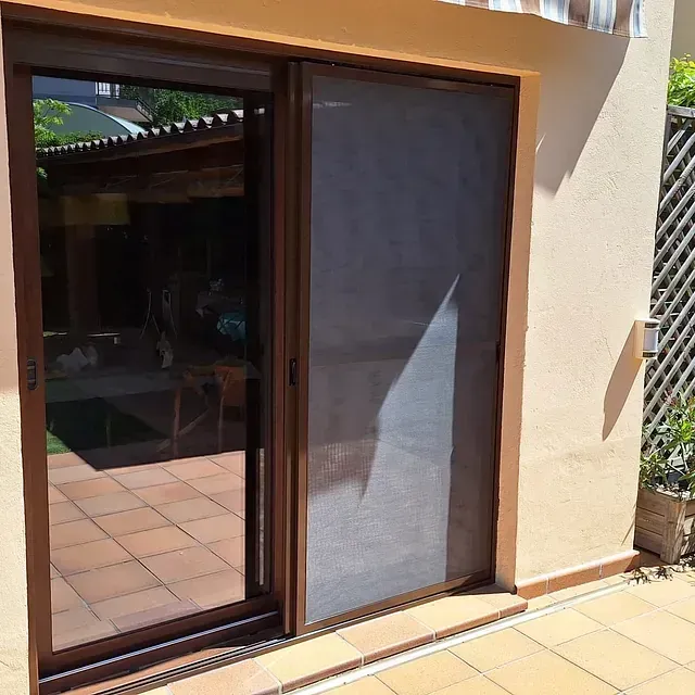 Puerta corrediza de vidrio con marco marrón y mosquitera. Da a un patio exterior con baldosas de terracota y pared beige.