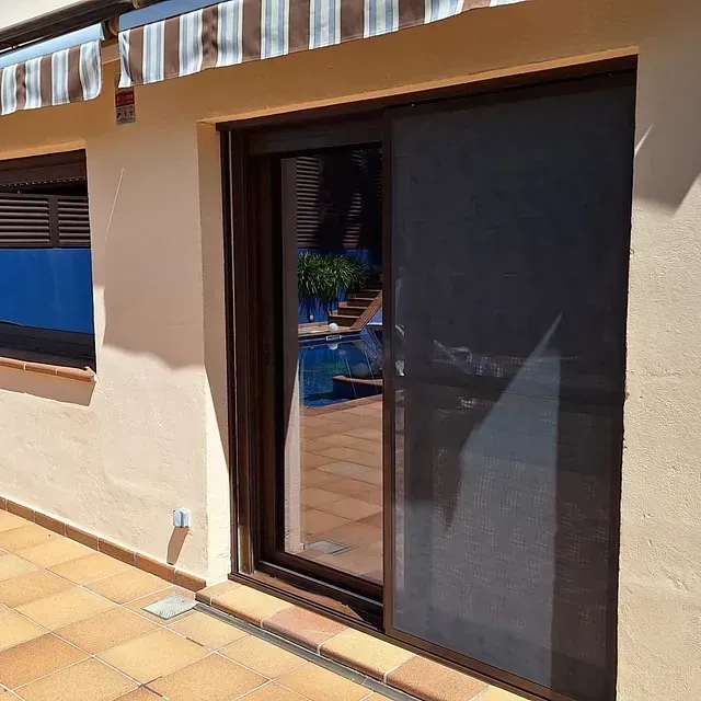 Una puerta corrediza de vidrio con marco marrón y mosquitera oscura. Se ve una piscina a través del cristal.