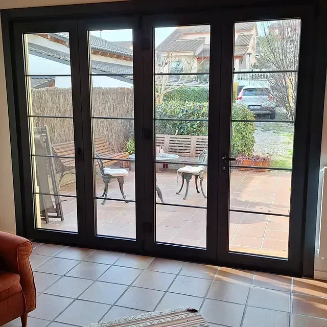 Una vista a través de puertas y ventanas de vidrio con marco oscuro hacia un patio con muebles; un automóvil está estacionado en una calle.