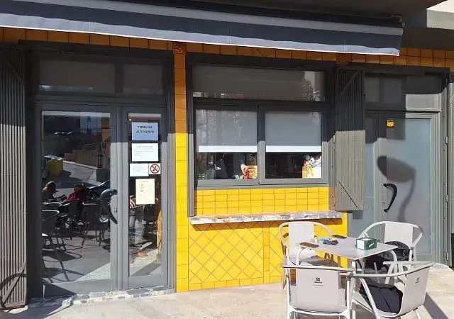 Exterior de un pequeño edificio con azulejos amarillos, toldo y mesas al aire libre. La gente se sienta en mesas, visibles a través de las ventanas.