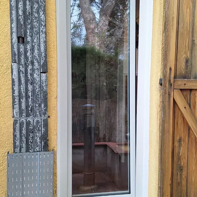 Una ventana alta y estrecha con marco blanco está flanqueada por una contraventana plegable de color gris desgastado y una puerta de madera.