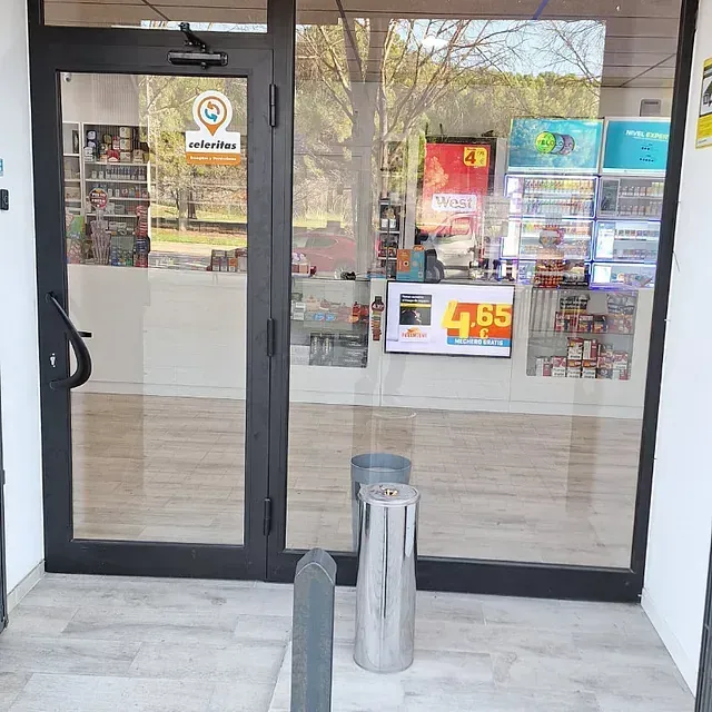 Entrada de una tienda con puertas de cristal. En el interior hay estanterías con productos, un expositor de precios y un cenicero.