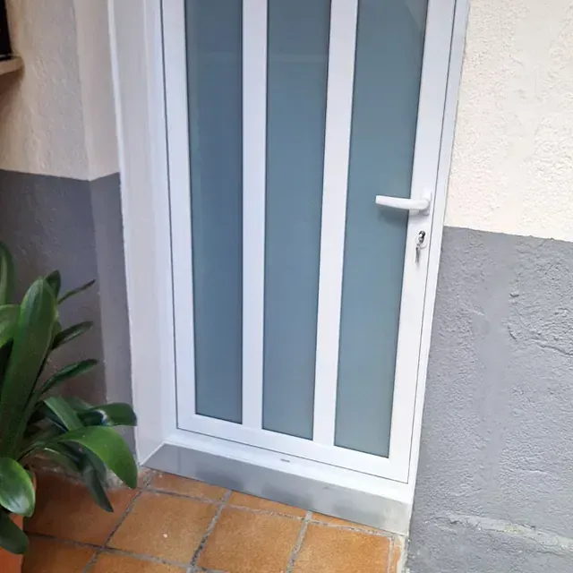 Puerta blanca con tres paneles de vidrio esmerilado. La puerta se encuentra en una pared con una combinación de pintura bitono gris y blanca; una planta y un suelo de terracota se encuentran en primer plano.