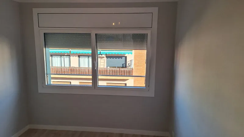 Una ventana en una habitación con vista a la fachada de un edificio, enmarcada por una moldura blanca. Las paredes son grises; la habitación está vacía.