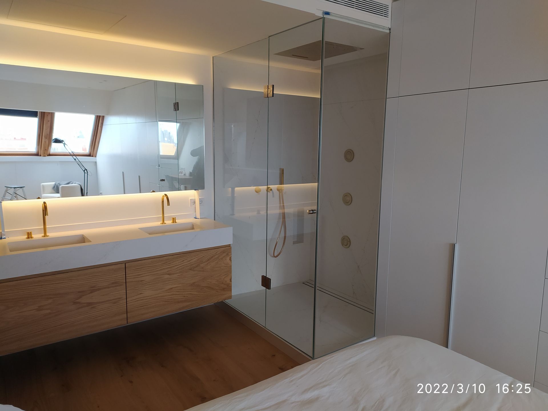 Un baño moderno con ducha con mampara de cristal, lavabo doble con grifería dorada y muebles de madera. Las paredes blancas y una claraboya iluminan el espacio.