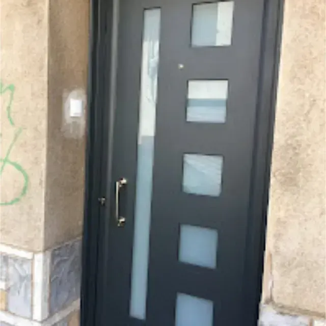 Una puerta de entrada gris con ventana vertical y paneles de vidrio cuadrados. La puerta tiene un tirador dorado y está empotrada en una pared color canela.