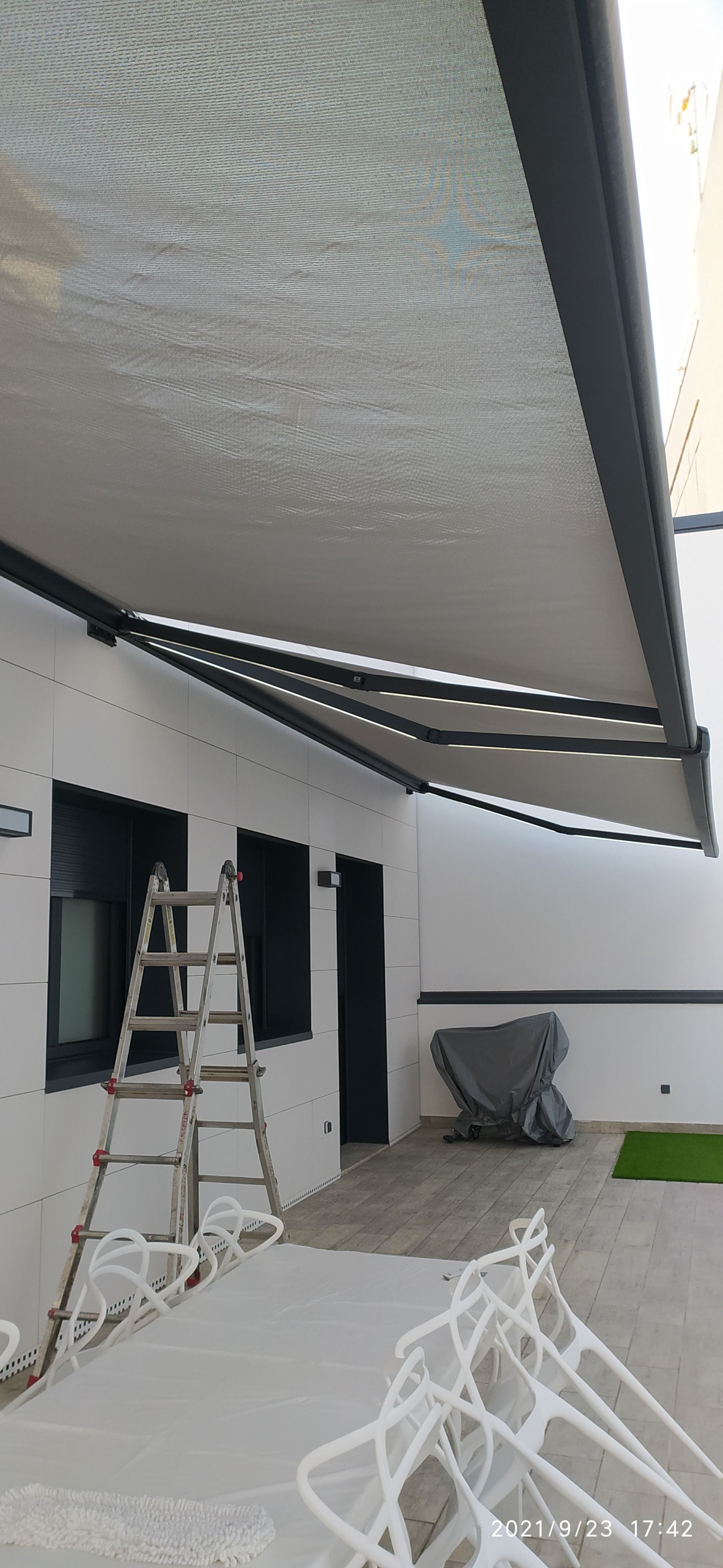 Un edificio blanco con toldos abiertos. Una escalera está apoyada contra la pared. Sillas blancas en primer plano.