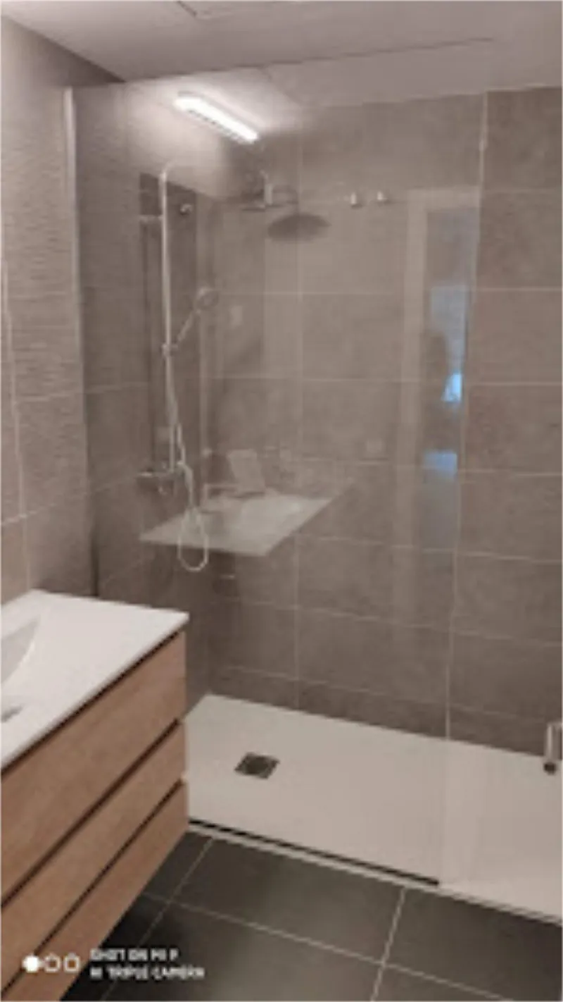 Baño moderno con mampara de ducha de cristal. Cuenta con paredes y suelo de azulejos grises, base de ducha blanca y tocador de madera.