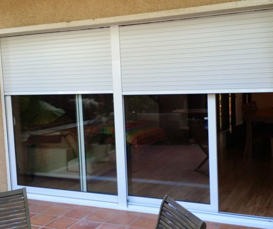 Puertas corredizas de vidrio blanco con persianas enrollables blancas cerradas, que reflejan un área de patio al aire libre y una habitación interior.