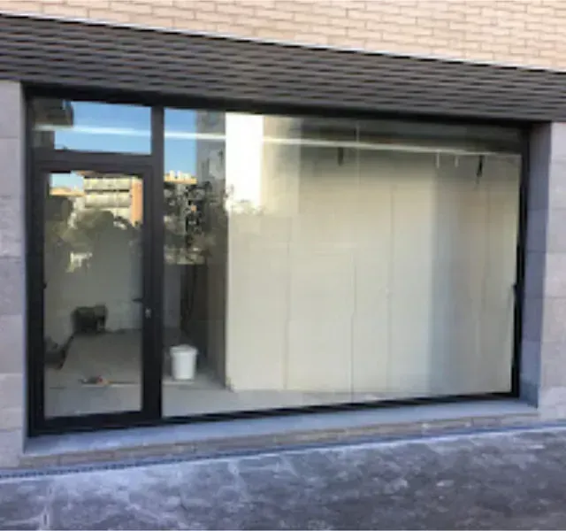 Escaparate vacío con ventanas de cristal y una puerta, marco oscuro, debajo de una fachada de ladrillo claro.