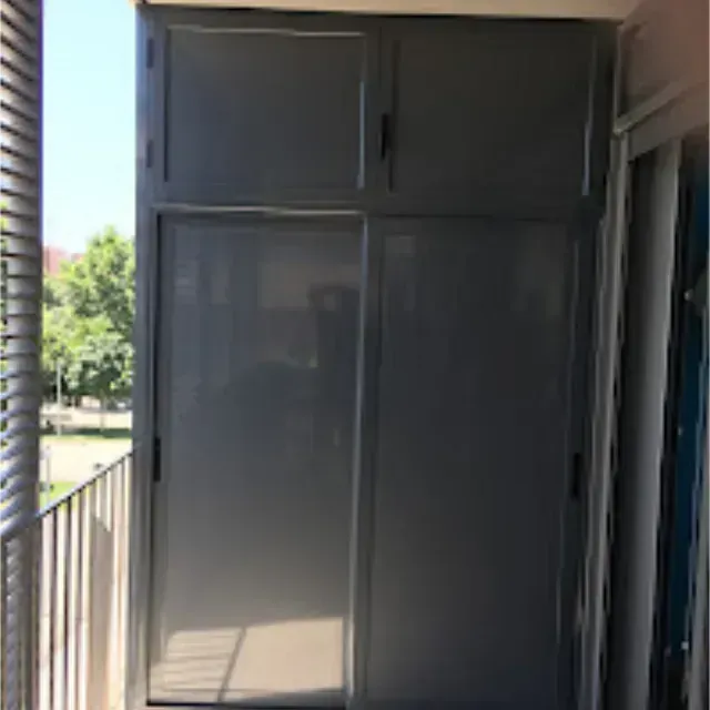 Mueble de almacenaje gris en un balcón, con dos puertas correderas y armarios superiores. La luz del sol ilumina la escena.