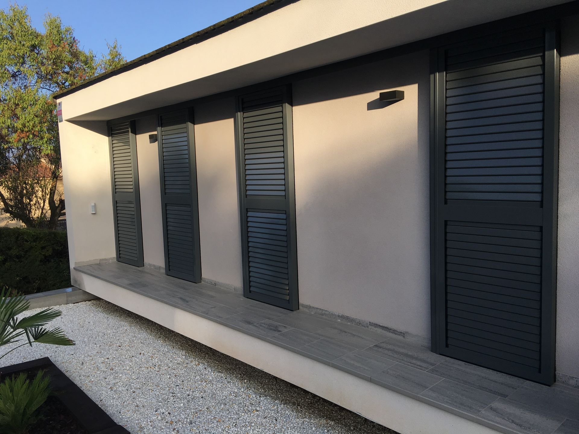 Exterior de un edificio moderno con persianas grises y paredes claras. Grava blanca y algo de vegetación en la fachada.