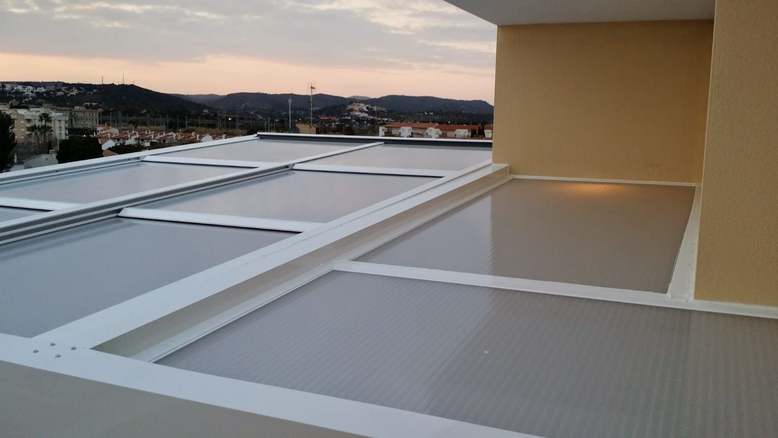 Vista exterior de un balcón con techo retráctil y opaco. Al fondo se aprecian paredes beige y un paisaje urbano.