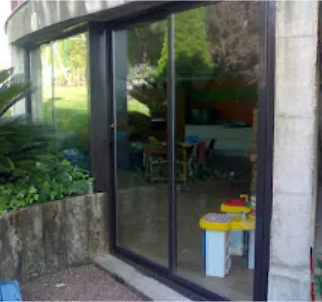 Puerta corrediza de vidrio que refleja un patio trasero y un interior con juguetes infantiles. El marco oscuro de la puerta contrasta con el exterior de piedra clara.