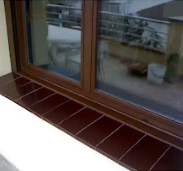 Primer plano de una ventana con marco marrón y azulejos marrones a lo largo del alféizar inferior, que refleja un balcón y el exterior.