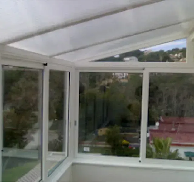 Vista interior de un solárium con marcos blancos y grandes ventanales con vistas a un paisaje verde y edificios con techos rojos.