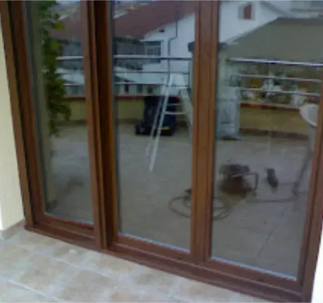 Puertas de vidrio con marco marrón que dan a un balcón. Las puertas reflejan un tejado, una escalera y herramientas.