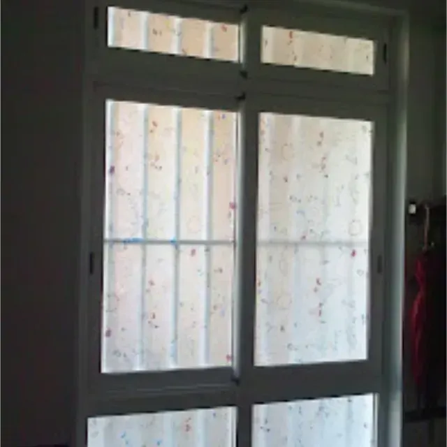 Ventana con marco blanco, dos paneles grandes y pequeños paneles superiores, cubiertos por cortinas transparentes con estampado floral.