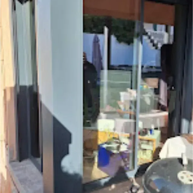 Una puerta corrediza de vidrio refleja una escena al aire libre con una mesa y una persona. El sol proyecta sombras en el marco de la puerta.