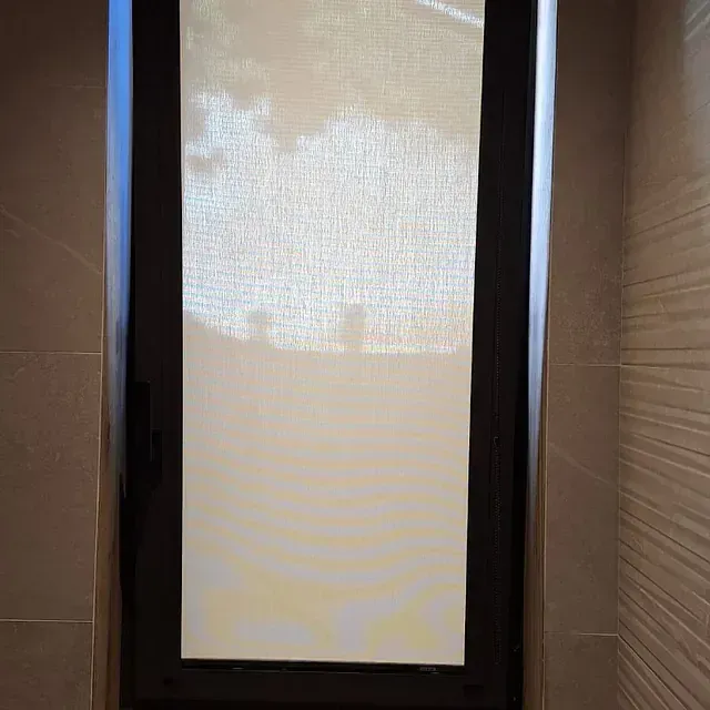 Ventana con una persiana enrollable cerrada de color claro dentro de un marco oscuro. La luz se filtra a través de la persiana. La ventana está empotrada en una pared de azulejos.