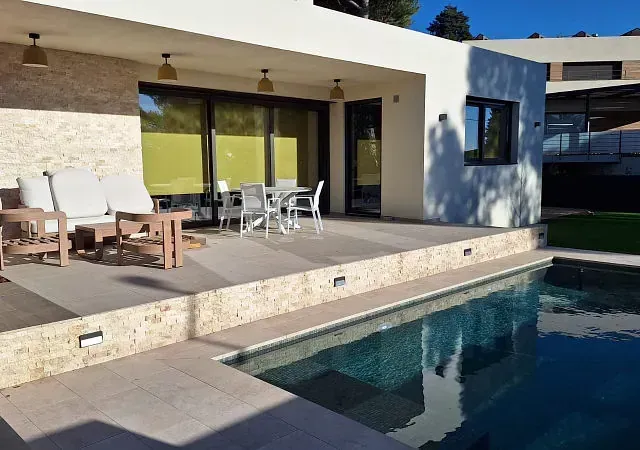 Una casa moderna con piscina. Cuenta con un patio exterior con asientos y mesa de comedor. El agua de la piscina refleja el cielo azul.