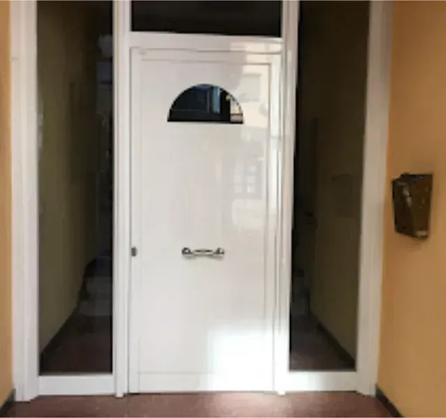 Puerta de entrada blanca con una pequeña ventana arqueada, ubicada en un pasillo beige con escaleras y un buzón.