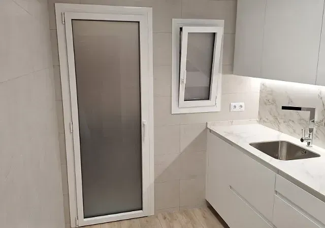 Cocina blanca con puerta de vidrio esmerilado, una pequeña ventana inclinada y gabinetes y encimeras blancos.