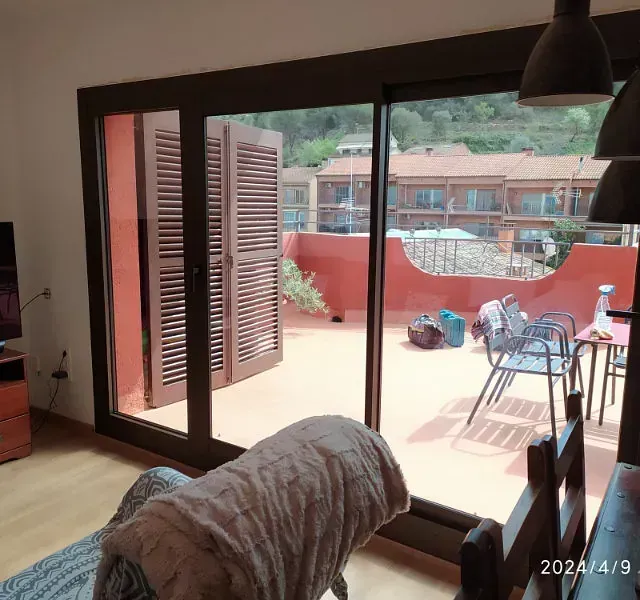 Vista interior de una habitación con una puerta corrediza de vidrio que conduce a una terraza en la azotea con tejas rojas con vista a edificios y vegetación.