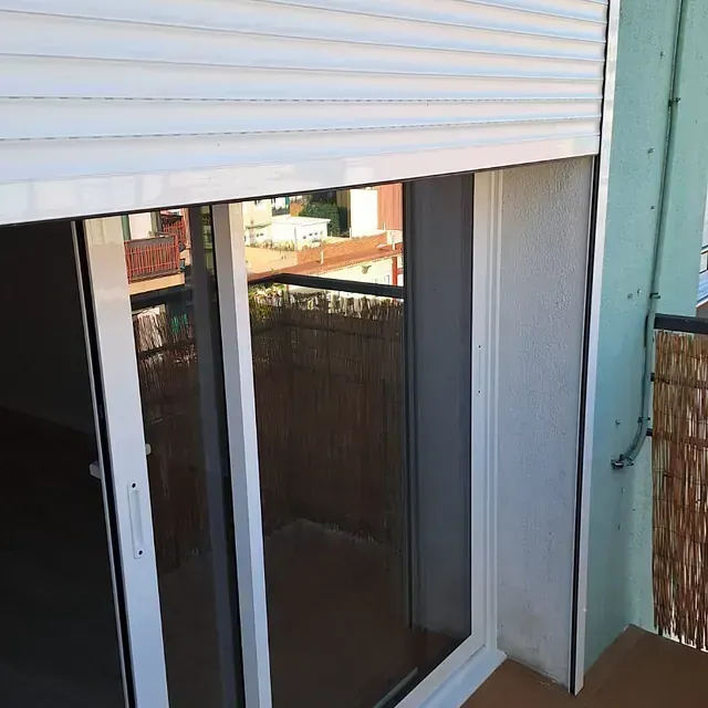 Una puerta corredera blanca con una persiana blanca parcialmente cerrada. La vista incluye un balcón y los edificios circundantes.