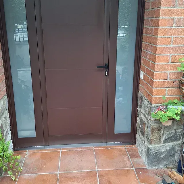 Puerta marrón con ventanales, rodeada de ladrillo y piedra, con suelo de baldosas de terracota.