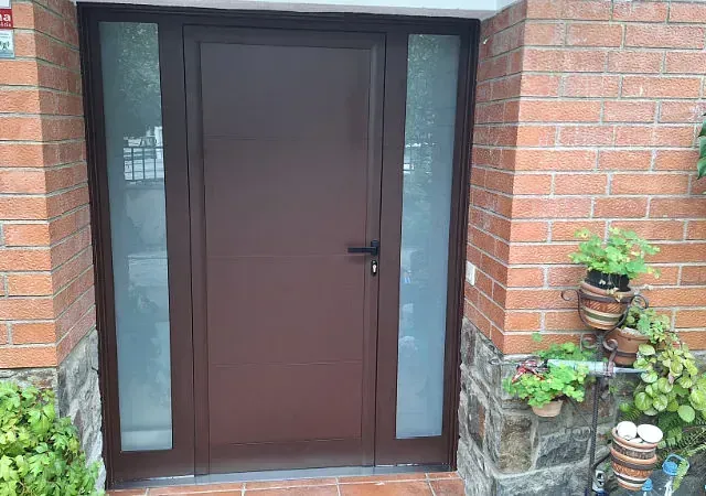Puerta marrón con laterales de vidrio esmerilado, empotrada en un edificio de ladrillo. Cerca hay macetas con plantas.
