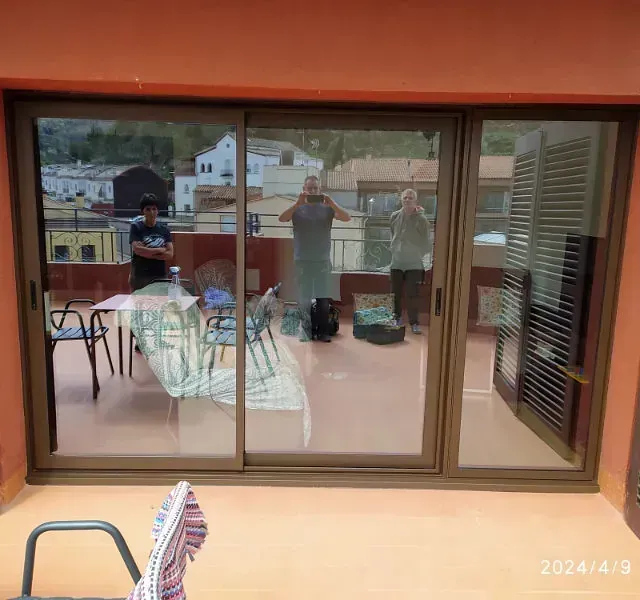 Tres personas en una terraza vistas a través de una puerta corrediza de cristal. Dos personas están de pie con los brazos en alto; una observa con los brazos cruzados. La escena incluye mesas, sillas y edificios al fondo.