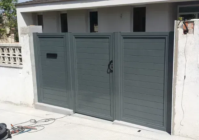 Tres portones metálicos grises, uno de ellos con buzón, frente a un edificio de color claro. Acera de hormigón visible.