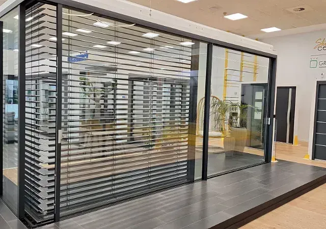 Exhibición de un sistema de puerta corredera de vidrio de gran tamaño con persianas integradas en una sala de exposición. La puerta tiene un marco oscuro y se encuentra sobre una plataforma elevada.