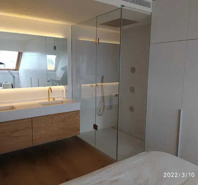 Baño moderno con ducha con mampara de cristal, lavabo blanco y muebles de madera. El espacio es luminoso y minimalista.