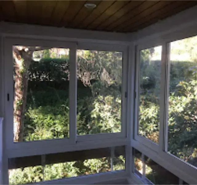 Un solárium con grandes ventanales con marcos blancos que ofrecen vistas a la exuberante vegetación de los árboles. El techo es de madera.