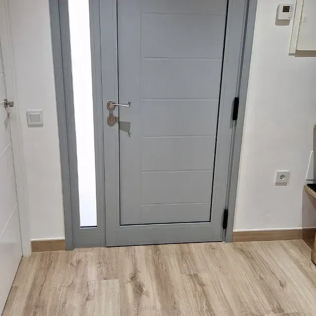 Una puerta interior gris con un panel de ventana, ubicada en una habitación de paredes blancas y piso con apariencia de madera clara.
