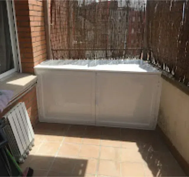 Gabinete blanco en un balcón iluminado por el sol con piso de baldosas, una cerca de caña y paredes de ladrillo.