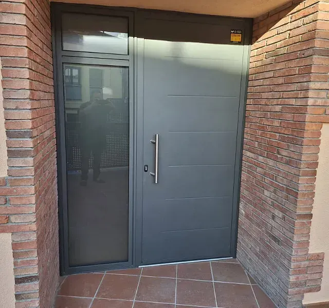 Puerta de entrada gris con ventana lateral y paredes de ladrillo. La puerta tiene líneas horizontales y un tirador plateado.